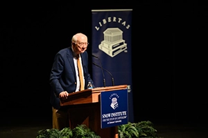 An older man speaks at a podium on a stage with a “Liberty” banner and a “Snow Institute” sign, with ferns in front.