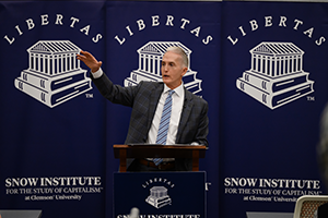 A speaker gestures while addressing an audience at the Snow Institute for the Study of Capitalism, with Libertas logos displayed in the background.