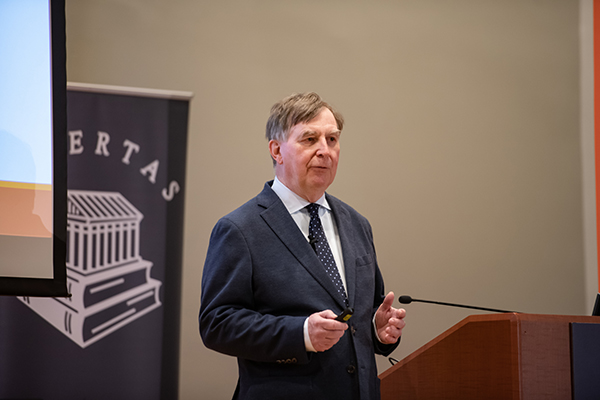 A speaker presenting at a conference, gesturing while discussing a topic related to education or academia.