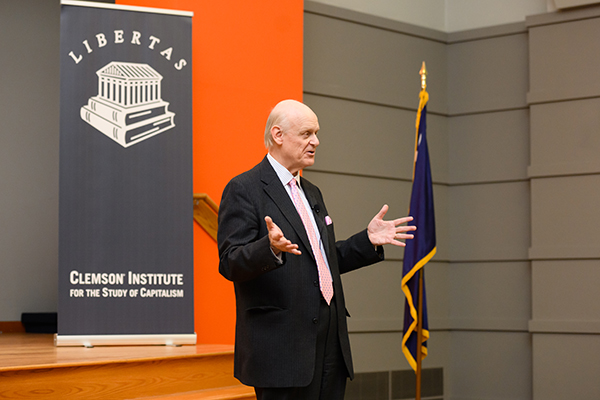 A speaker presenting at the Clemson Institute for the Study of Capitalism, with a Libertas banner in the background.