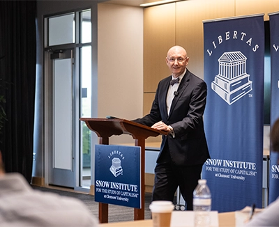 Man in a suit speaks at a podium during a Snow Institute event, with Libertas and Snow Institute banners behind him.