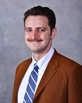A professional headshot of a man wearing a brown blazer and a blue and white striped tie, smiling confidently against a muted background.
