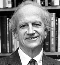 Black and white portrait of a smiling man with gray hair, wearing a suit and tie, set against a bookshelf backdrop.