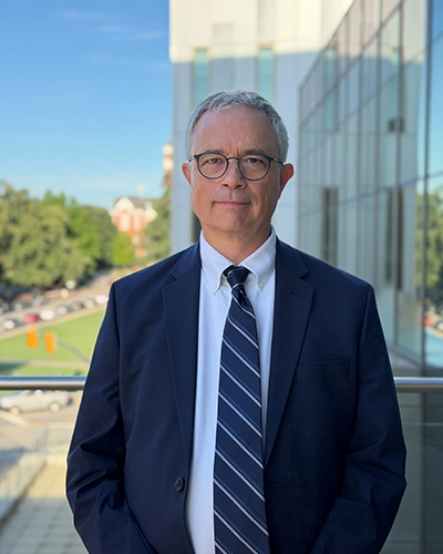 Professional head-and-torso portrait of a man in a navy suit and striped tie standing outdoors beside a modern glass building.