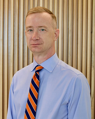 Professional portrait of a man with short hair wearing a light blue dress shirt and a striped orange and blue tie, posed against a wooden slat background.