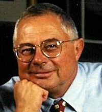 A close-up portrait of a smiling older man with glasses and a blue shirt.