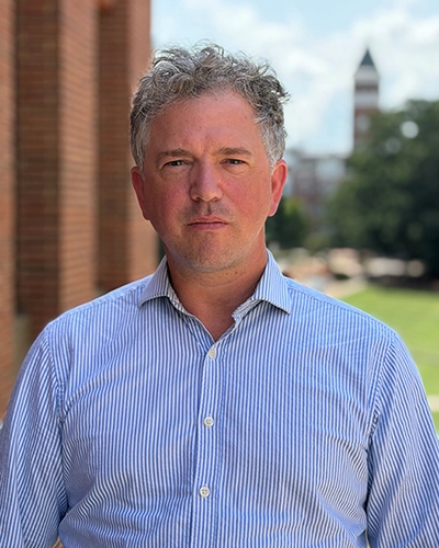 Portrait of a man in a light blue striped button-down shirt standing outdoors, with a brick building and a distant tower blurred in the background.