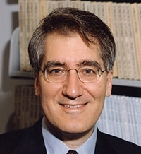 Headshot of a smiling man with glasses and short brown hair, dressed in a suit, set against a background of books or documents.