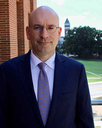 A professional portrait of a man in a suit with glasses, standing outdoors with a university building and green lawn in the background.