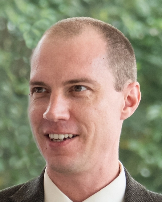 Portrait of a smiling man in a suit with short hair and a light background.
