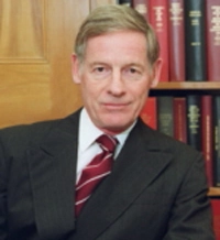Professional portrait of a man in a suit with a striped tie, sitting against a backdrop of bookshelves.