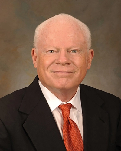 Portrait of a middle-aged man with white hair, smiling, wearing a black suit and orange tie.