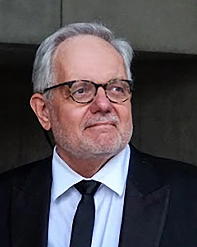 A smiling man in a tuxedo with glasses, standing against a neutral background.
