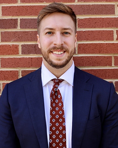 Professional headshot of a man in a suit with a patterned tie, standing in front of a brick wall.