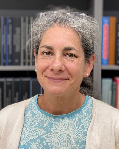 A smiling woman with gray hair, wearing a blue patterned shirt and a light cardigan, stands in front of a bookshelf filled with various books.
