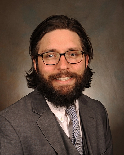 Professional portrait of a man with a beard, wearing glasses and a gray suit.
