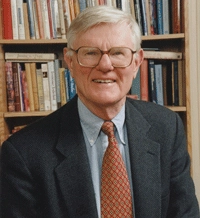 A smiling elderly man with glasses wearing a suit and tie, standing in front of a bookshelf filled with books.