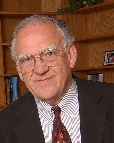 Portrait of an older man in a suit and tie, smiling warmly against a bookshelf background.
