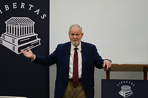 A speaker gesturing while presenting at a conference, with a "Libertas" logo featuring books and a classical building in the background.