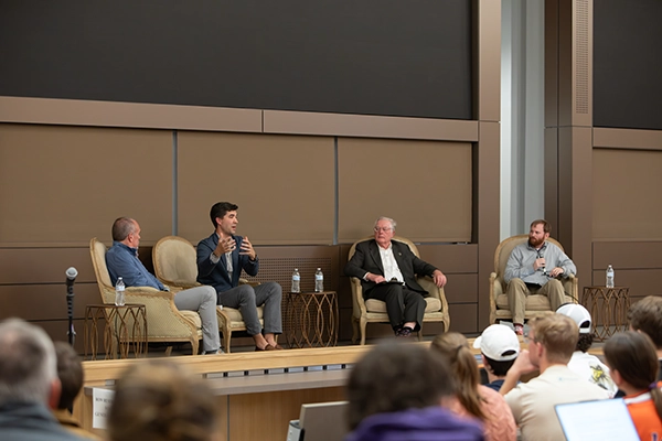 A panel discussion featuring four speakers seated on a stage, engaging with an audience in a lecture hall setting.