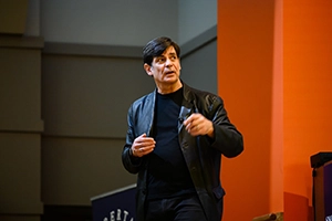 Speaker in a dark jacket gestures while presenting on stage in front of an orange wall.