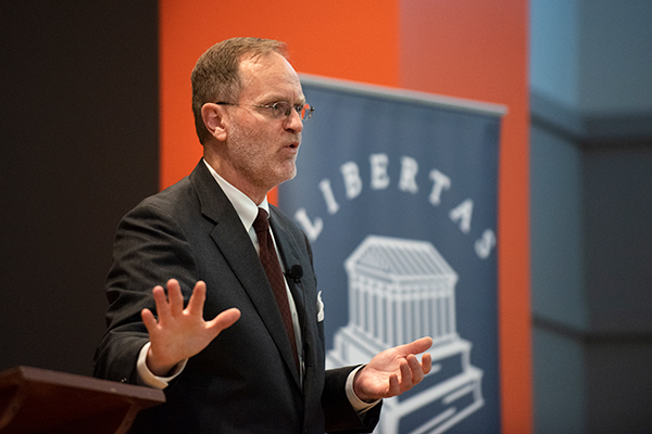A speaker passionately presents at a podium, gesturing with hands, in front of a banner featuring the word "Libertas."