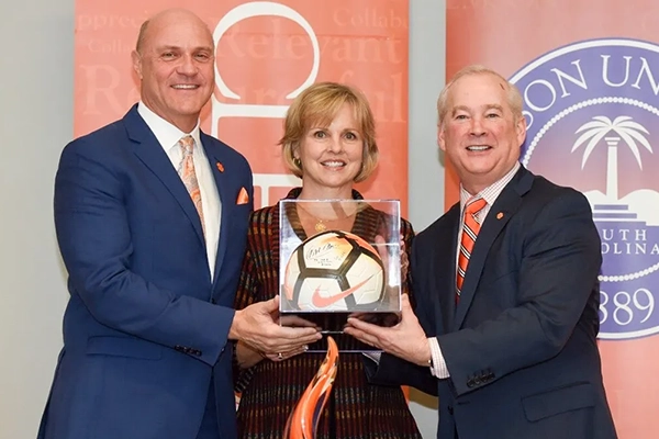 Three individuals standing together, presenting an award featuring a soccer ball in a clear display case. The backdrop includes banners with the text "Relevant," "Collaborate," and "Mission Un."