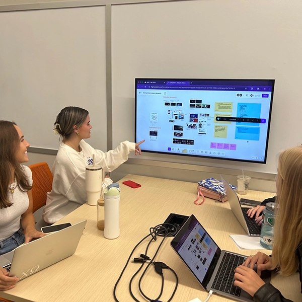 A group of three students collaborating in a meeting room while viewing a presentation on a large screen.