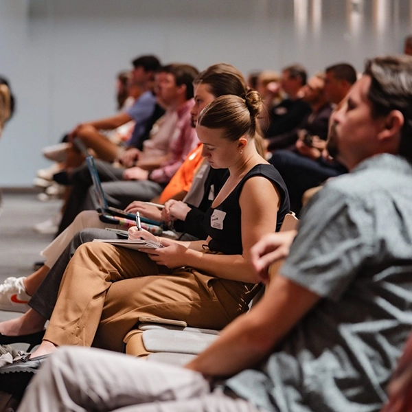 Audience members seated in a conference setting, focused on taking notes and engaging with the presentation.