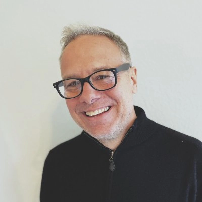 Professional headshot of a smiling man with glasses and short, light hair, wearing a black zip-up sweater against a neutral background.