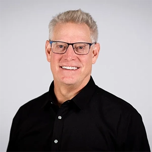 Headshot of a smiling man with short light-colored hair, wearing glasses and a black shirt.