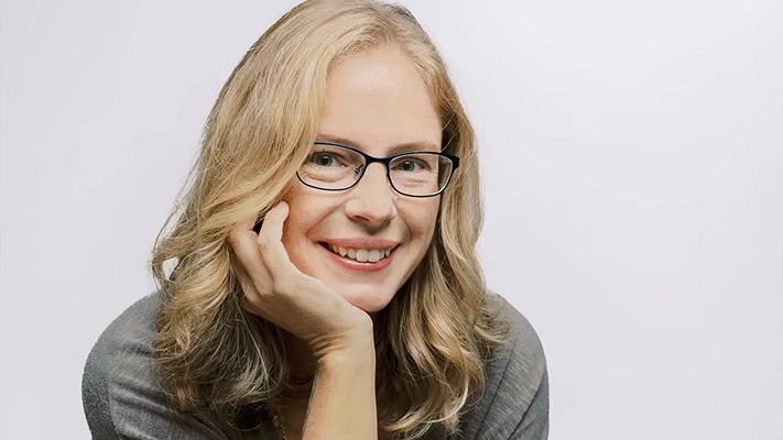 A woman with blonde, wavy hair and glasses smiles while resting her chin on her hand.