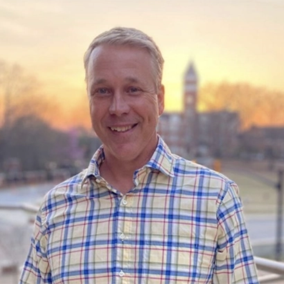 A smiling man wearing a plaid shirt with a soft sunset backdrop.