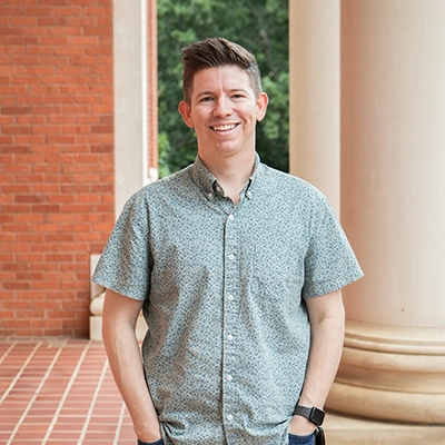 Smiling man in a short-sleeved patterned shirt standing outdoors near brick columns.