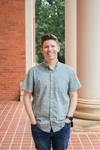 A smiling man stands confidently in a casual outfit, framed by columns in an outdoor setting.