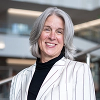 Smiling woman with long gray hair wearing a striped blazer in a modern office setting.