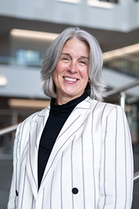 Portrait of a smiling woman with gray hair, wearing a striped blazer and a black turtleneck, standing indoors.
