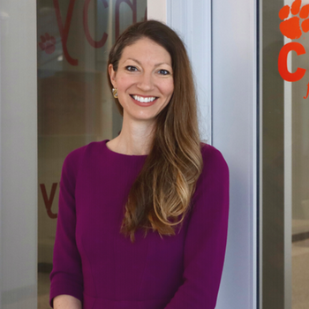 Smiling woman in a purple dress stands in front of a glass door with a Clemson University logo.