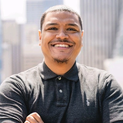Smiling man in a grey polo shirt poses confidently with arms crossed against an urban backdrop.