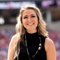 A smiling woman wearing a black sleeveless top and accessories with a blurred stadium background.