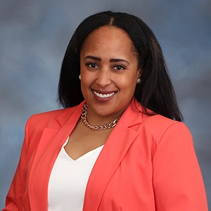 A professional portrait of a woman in a coral blazer, smiling confidently against a blue-gray background.