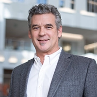 A professionally dressed man with gray hair and stubble, smiling in an office environment.