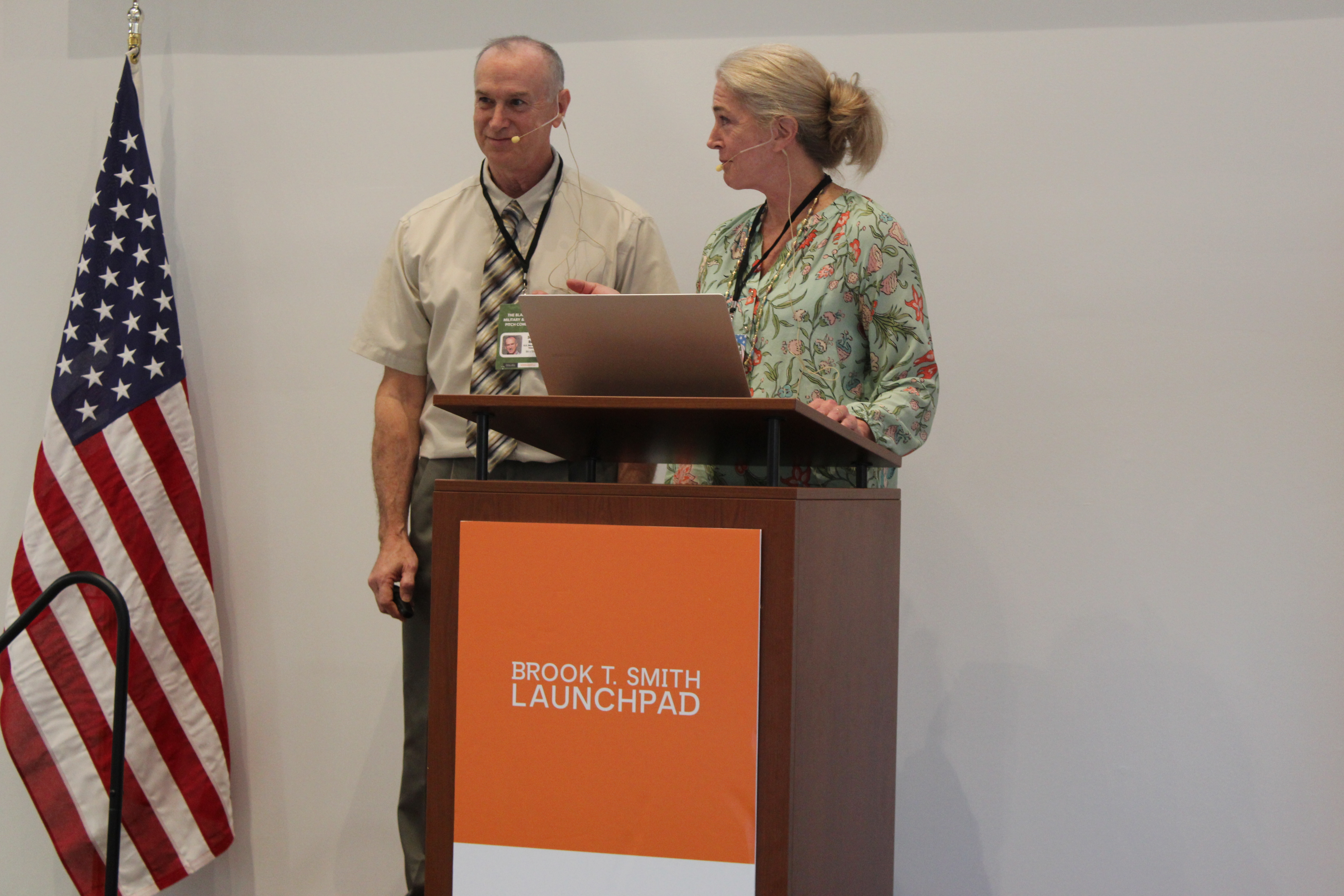 Two presenters wearing headsets stand at a podium with a laptop during a talk, with a U.S. flag nearby.