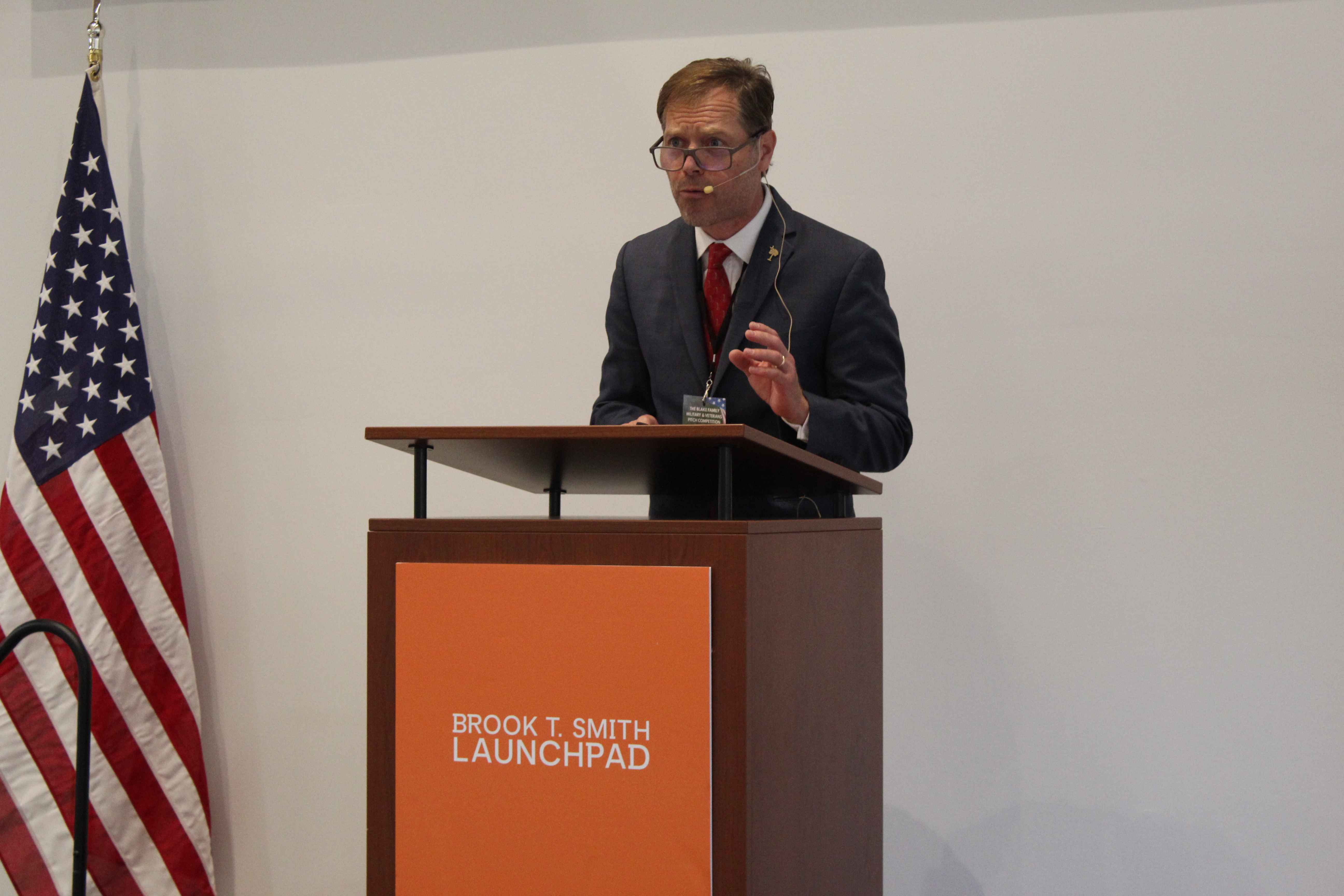Man speaking at a podium with a headset microphone, with an American flag beside him.