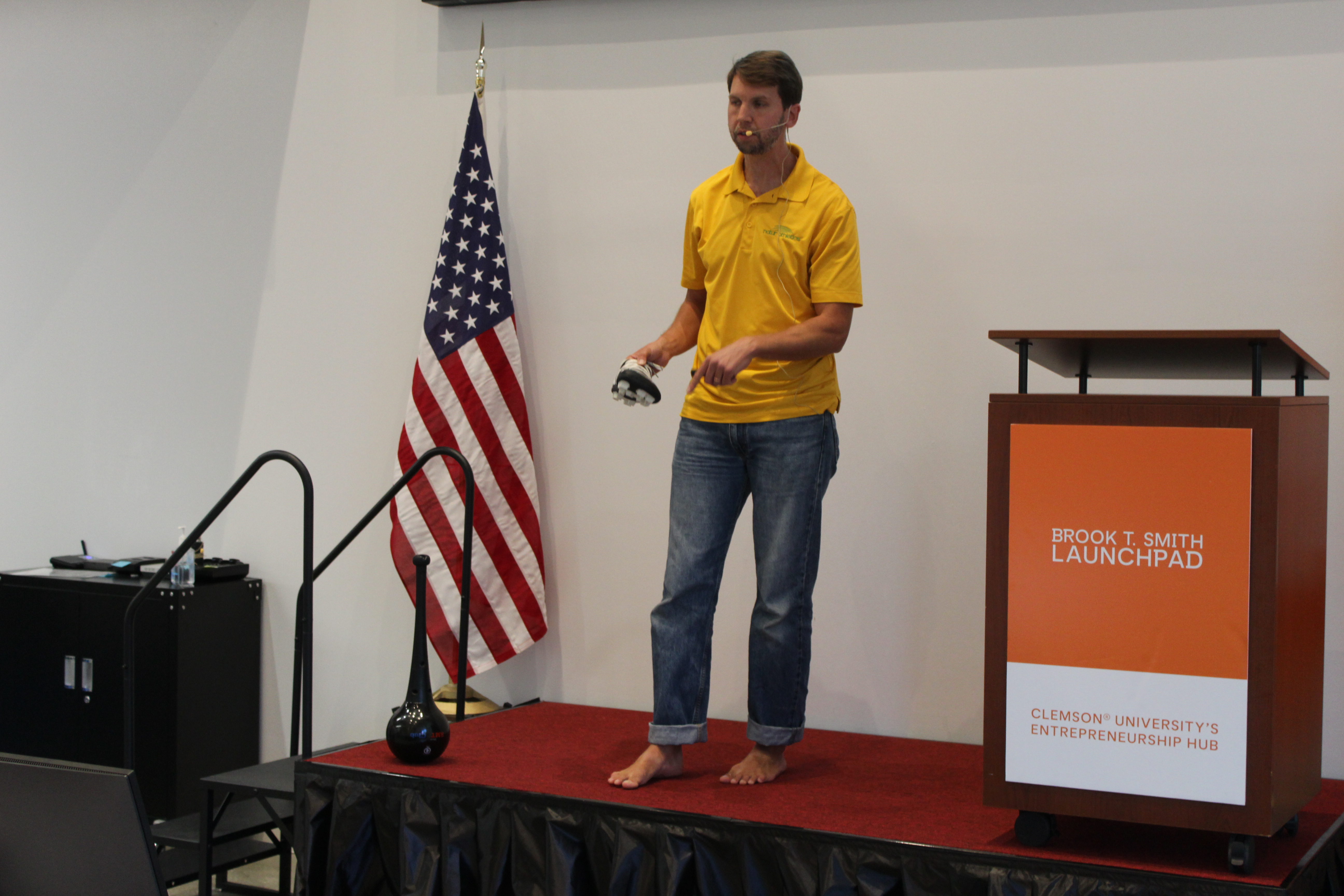 Man giving a presentation on a small stage, holding a device, with an American flag and a Launchpad podium nearby.