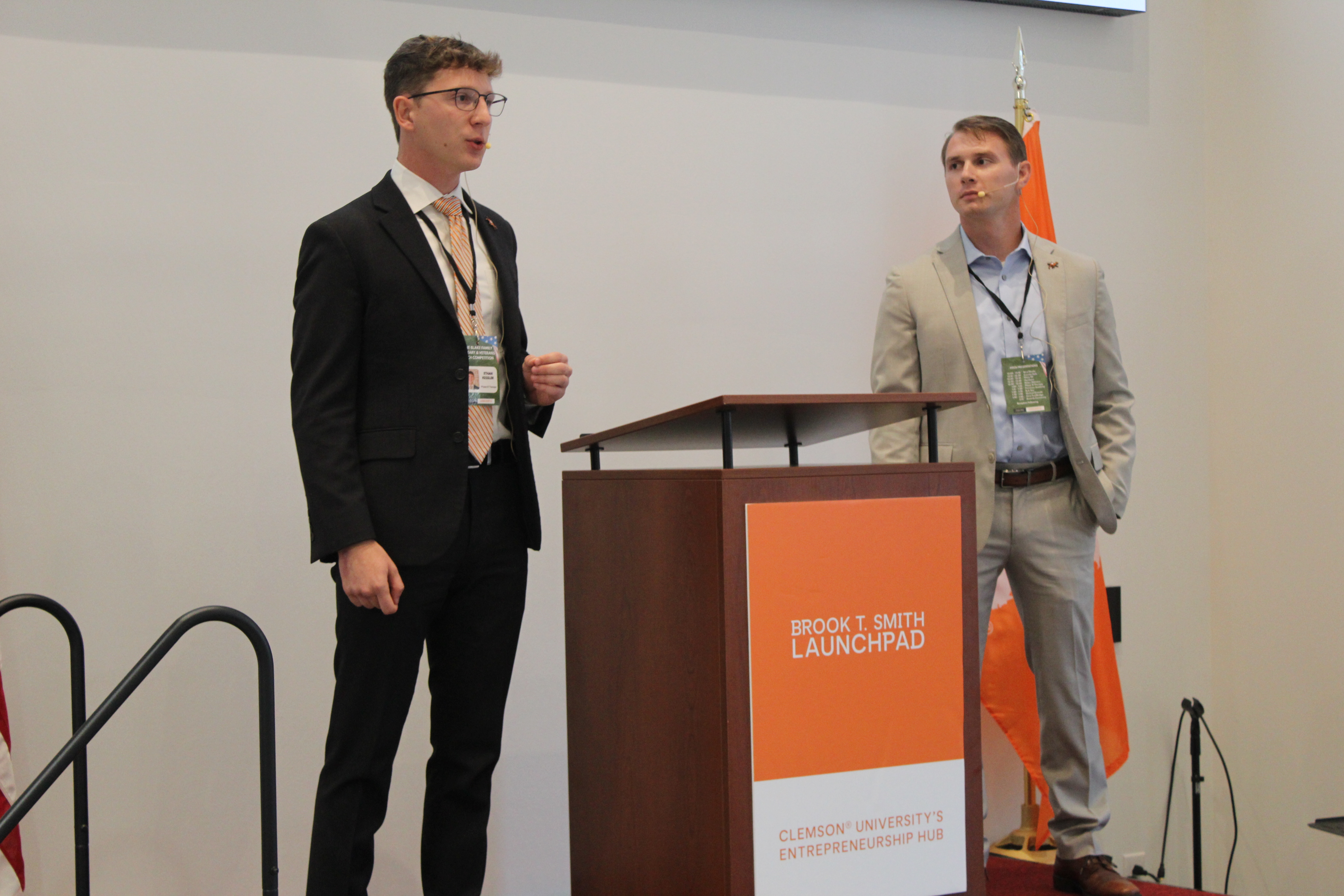 Two presenters in suits stand beside a podium at a Brook T. Smith Launchpad event, with one speaking into a headset microphone.