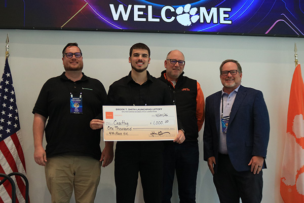 Award recipient and judges holding oversized checks beneath a large welcome banner with U.S. and Clemson flags beside them.