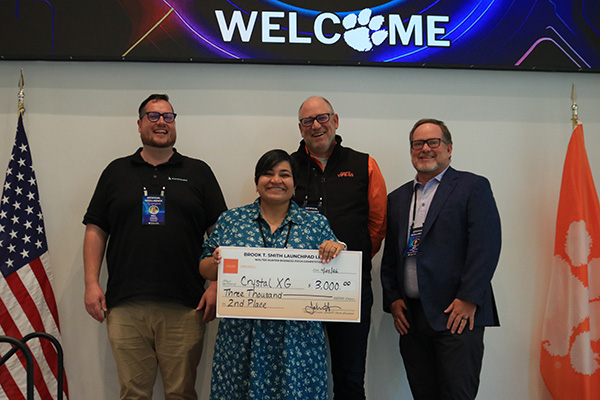 Award recipient and judges holding oversized checks beneath a large welcome banner with U.S. and Clemson flags beside them.