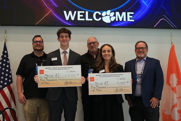 Award recipient and judges holding oversized checks beneath a large welcome banner with U.S. and Clemson flags beside them. 