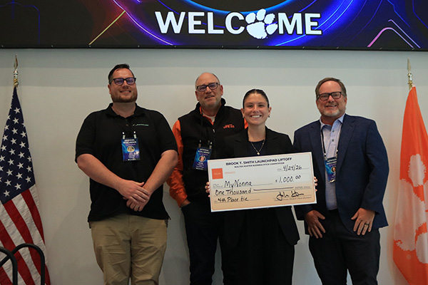 Award recipient and judges holding oversized checks beneath a large welcome banner with U.S. and Clemson flags beside them.